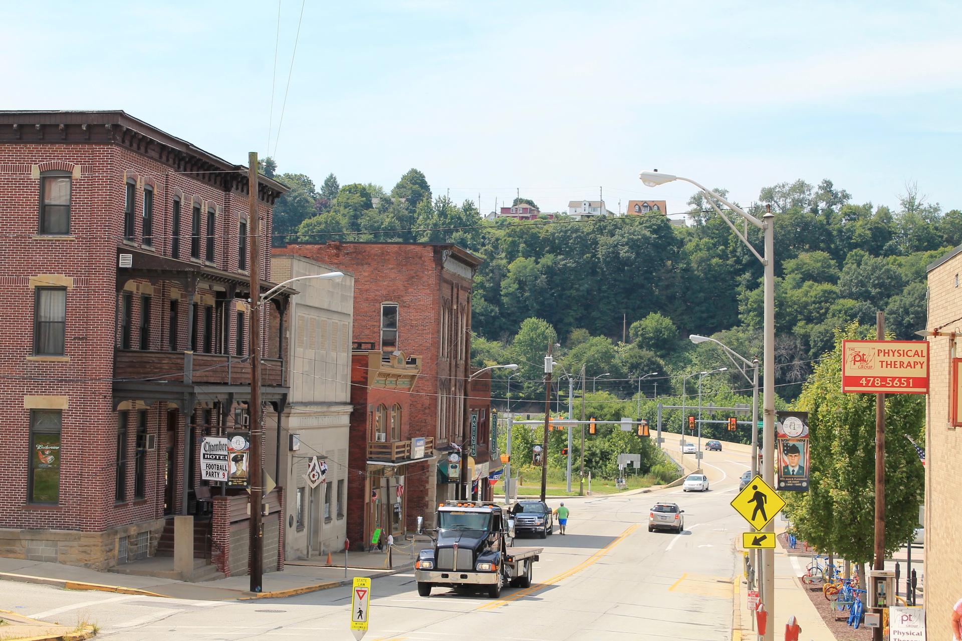 Busy street with brick buildings, cars, and distant green hills under daylight.