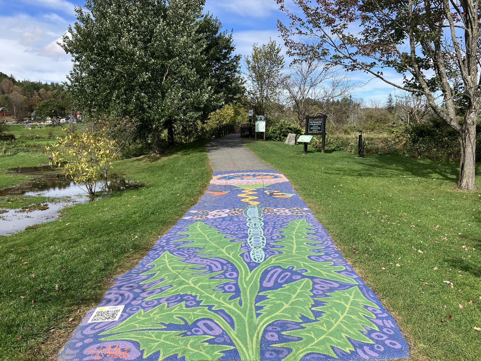 Paved path with a colorful mural, surrounded by green grass and trees.