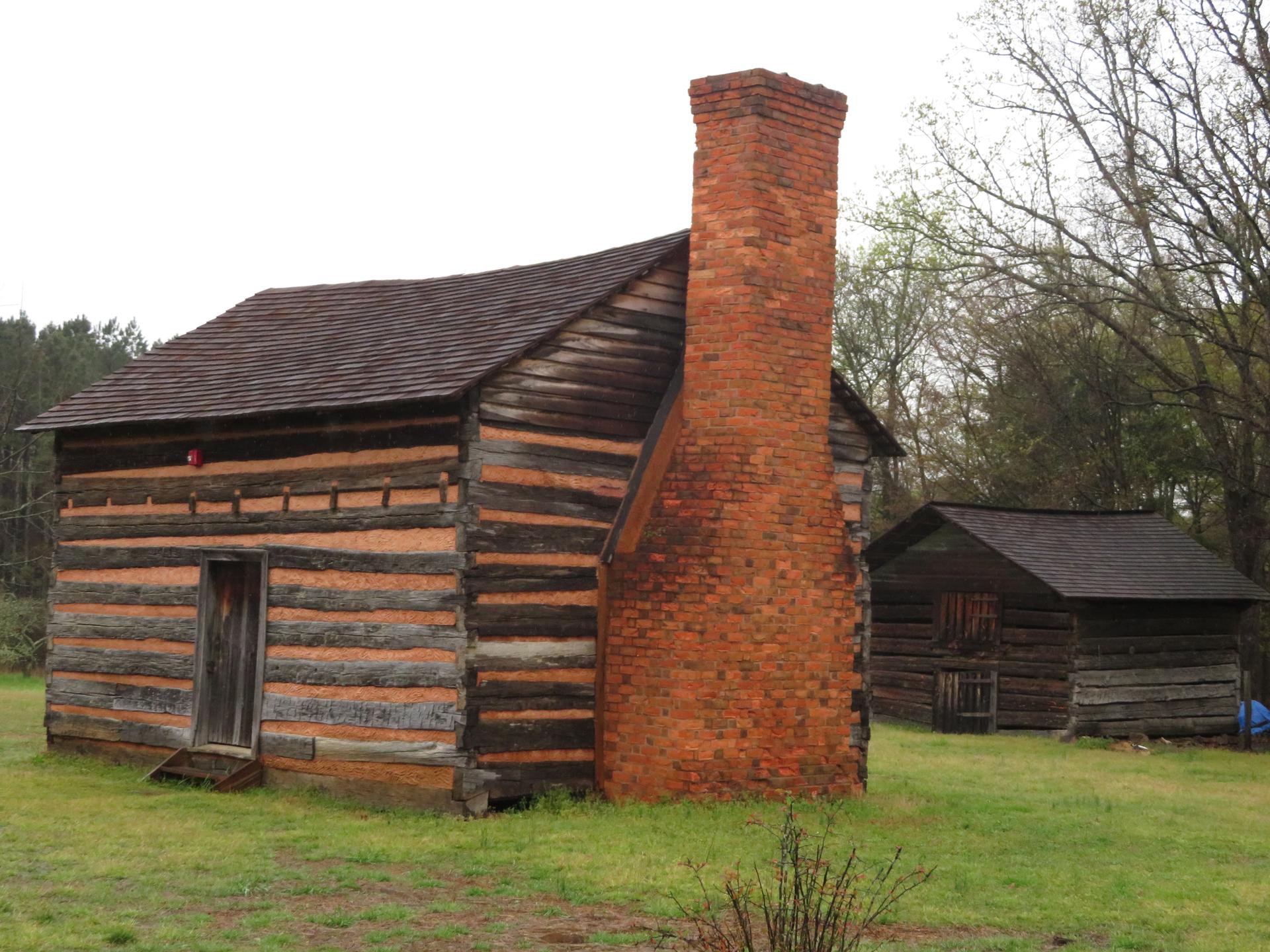 An old log cabin with a large brick chimney in a grassy field.