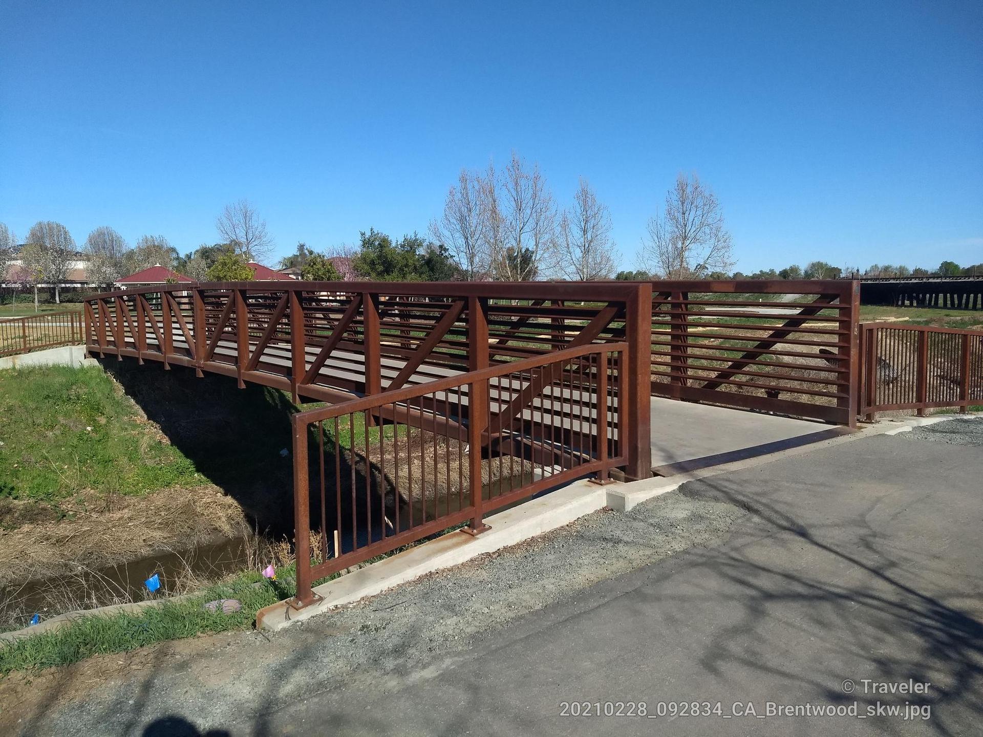 A brown metal pedestrian bridge spans a small ditch under a clear blue sky.