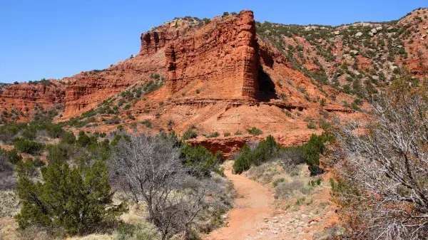Dirt path winds past a towering red rock formation under clear blue sky.