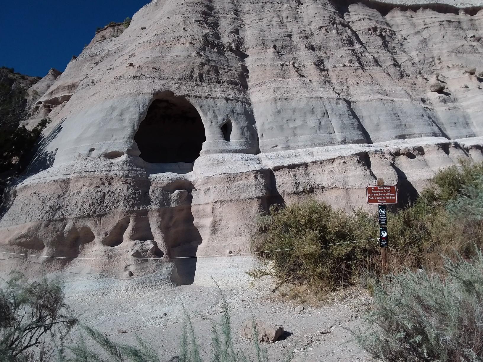 Large rock face with dark cave openings and a small sign, under bright light.