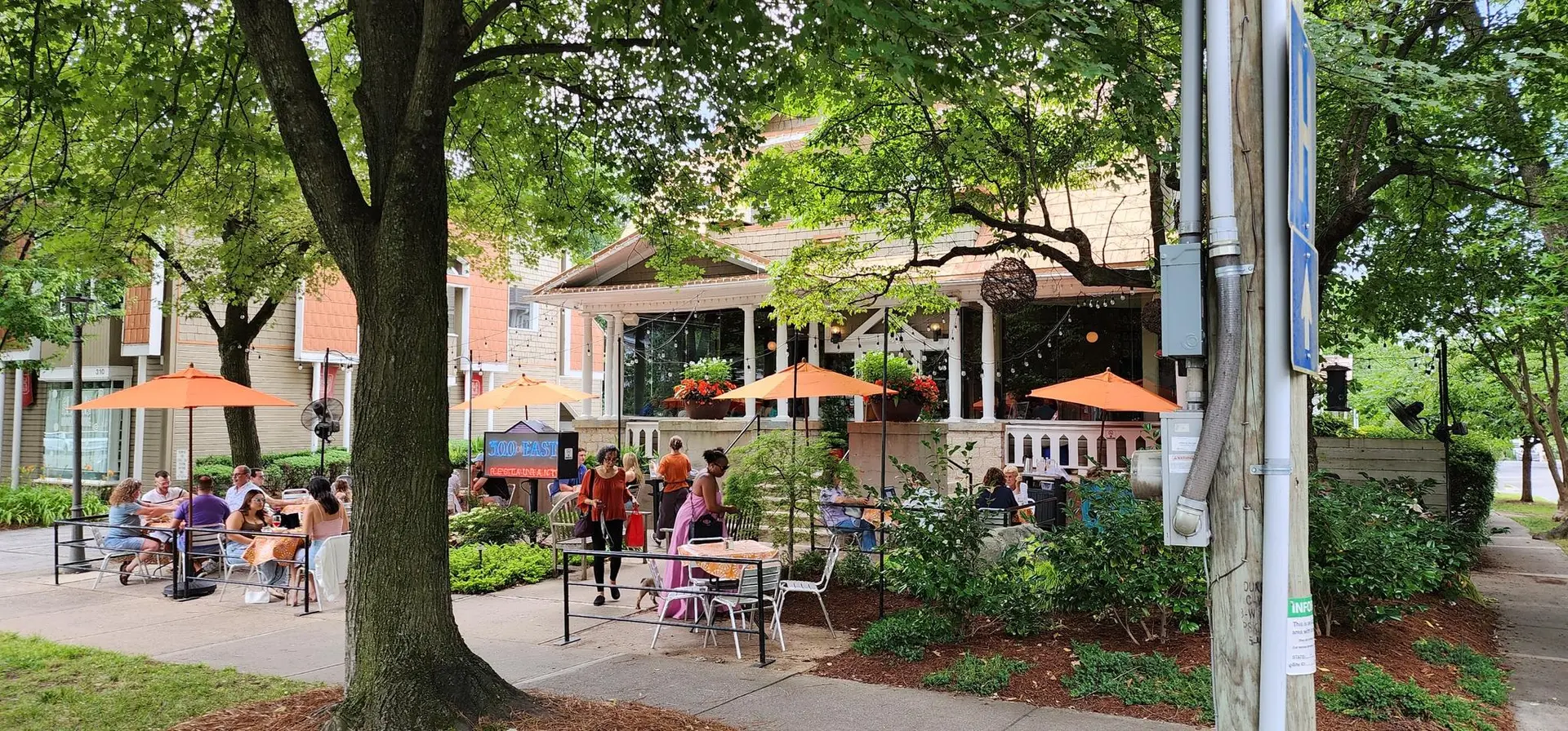 Shaded outdoor patio with people dining under orange umbrellas on a bright day.