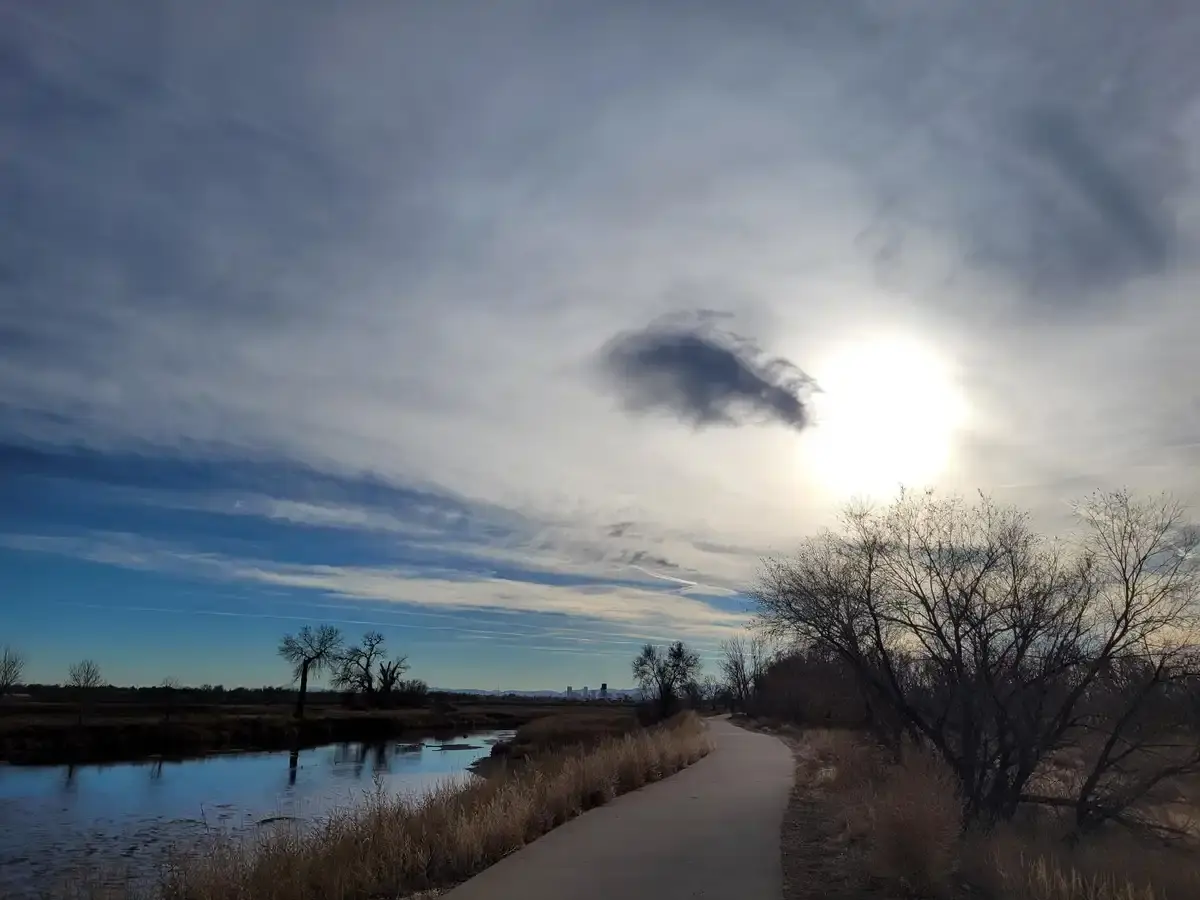 Paved path beside a river with bare trees under a bright, cloudy sky.
