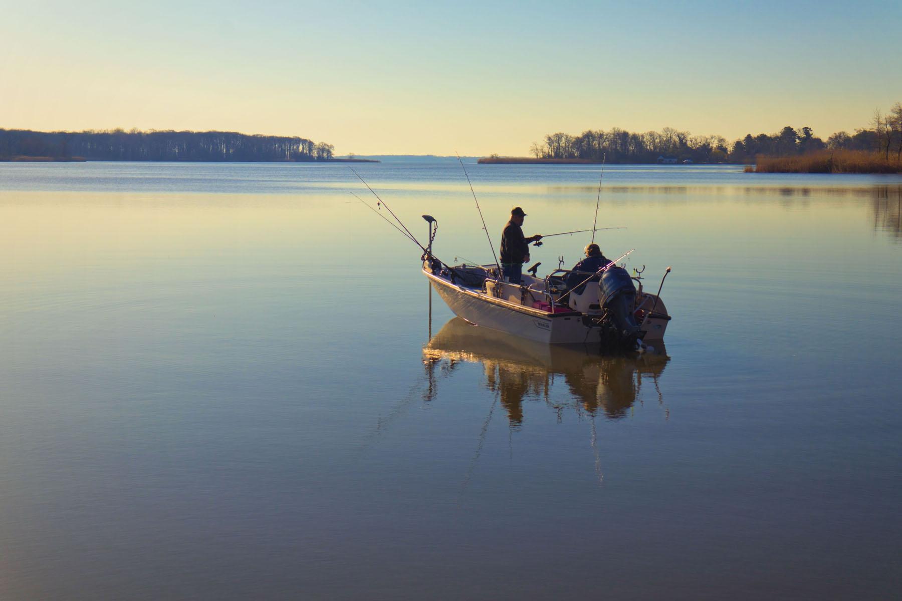 Two people fishing from a boat on calm water at sunset.