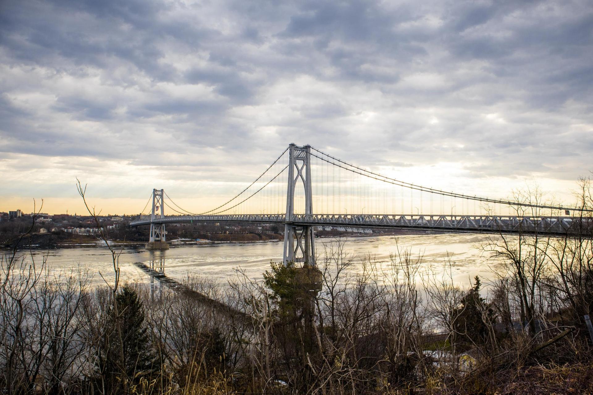Suspension bridge over a wide river under an overcast sky at dusk.