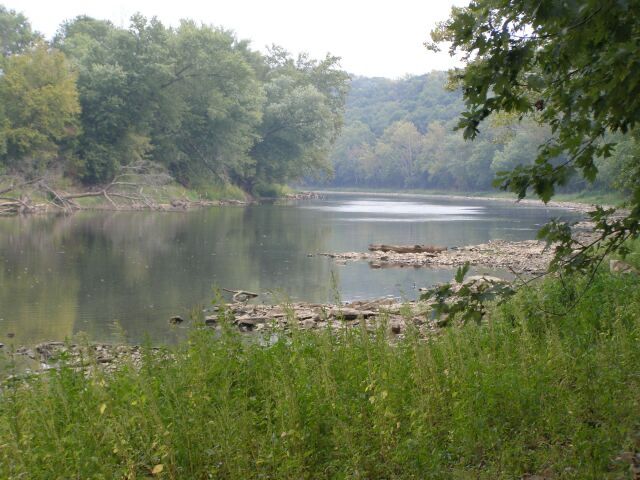 Calm river with rocky banks surrounded by dense green trees on an overcast day. - Milwaukee Road Transportation Trailway
