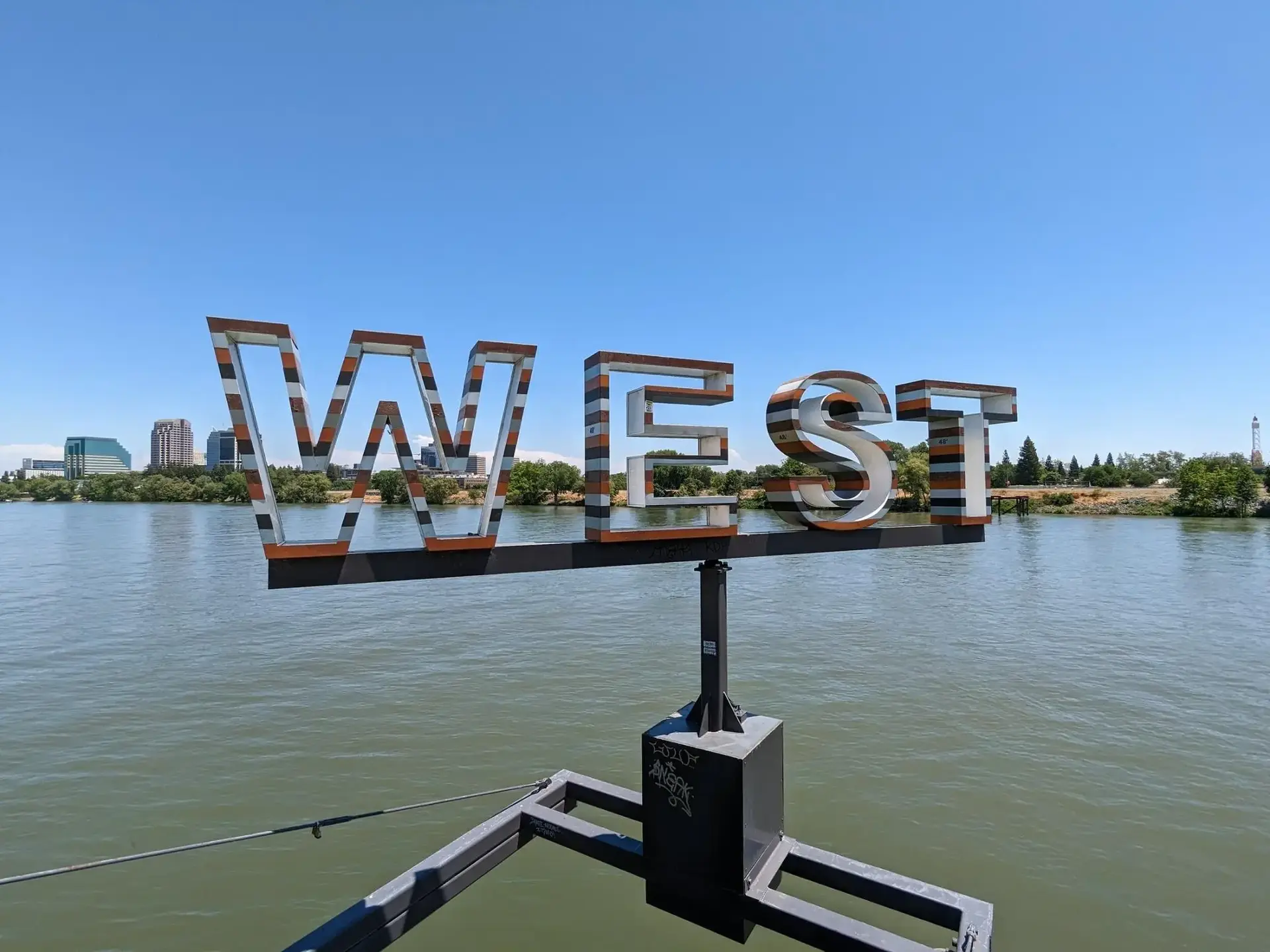 Large 'WEST' sign over water with a distant city skyline under clear skies.