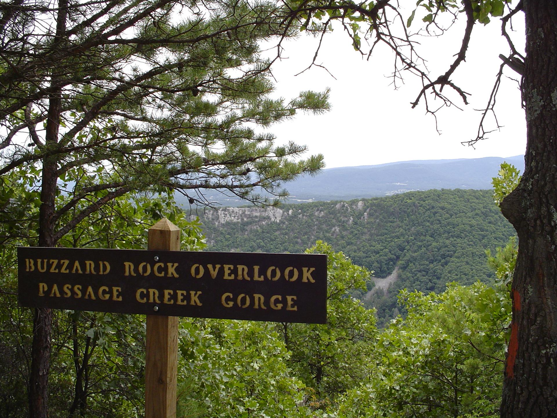 A wooden sign stands before a vast forested valley and distant cliffs.