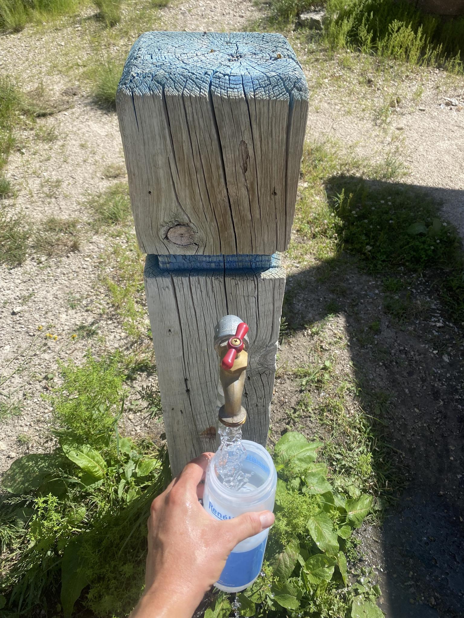 Hand filling a water bottle from a spigot on a wooden post in bright sunlight.