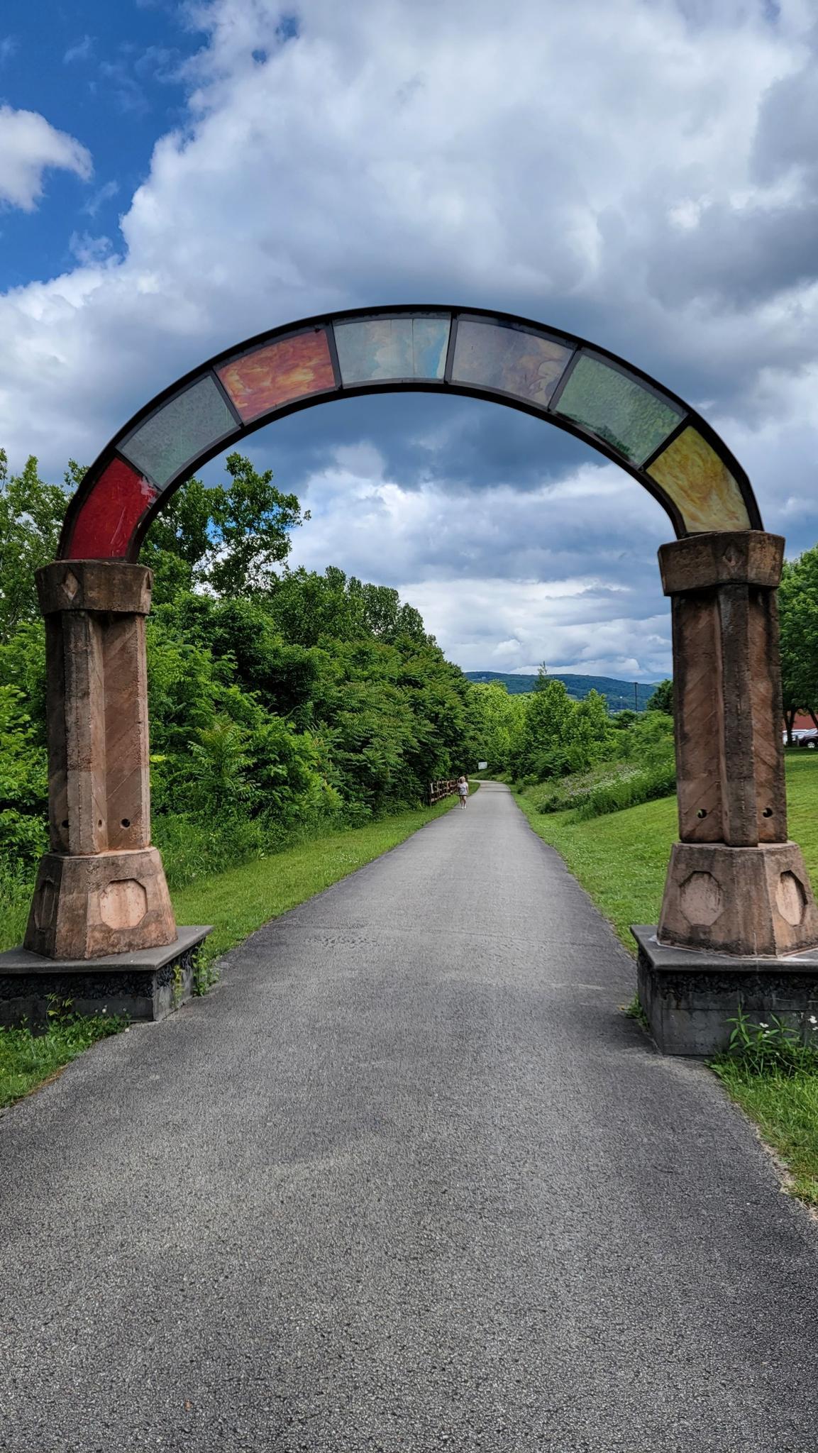 Colorful stained-glass arch over a paved path through green trees, cloudy sky.