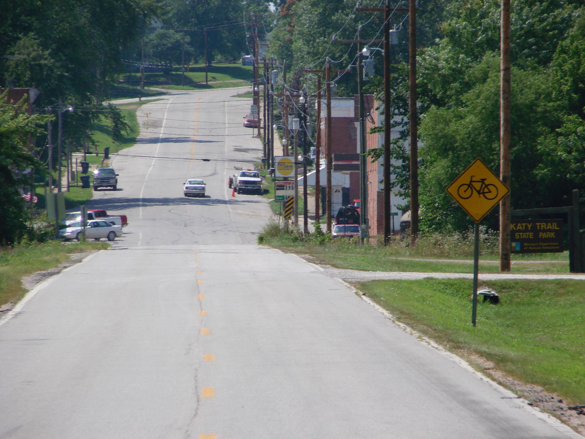 Uphill paved road with parked cars, power lines, and a bicycle sign.