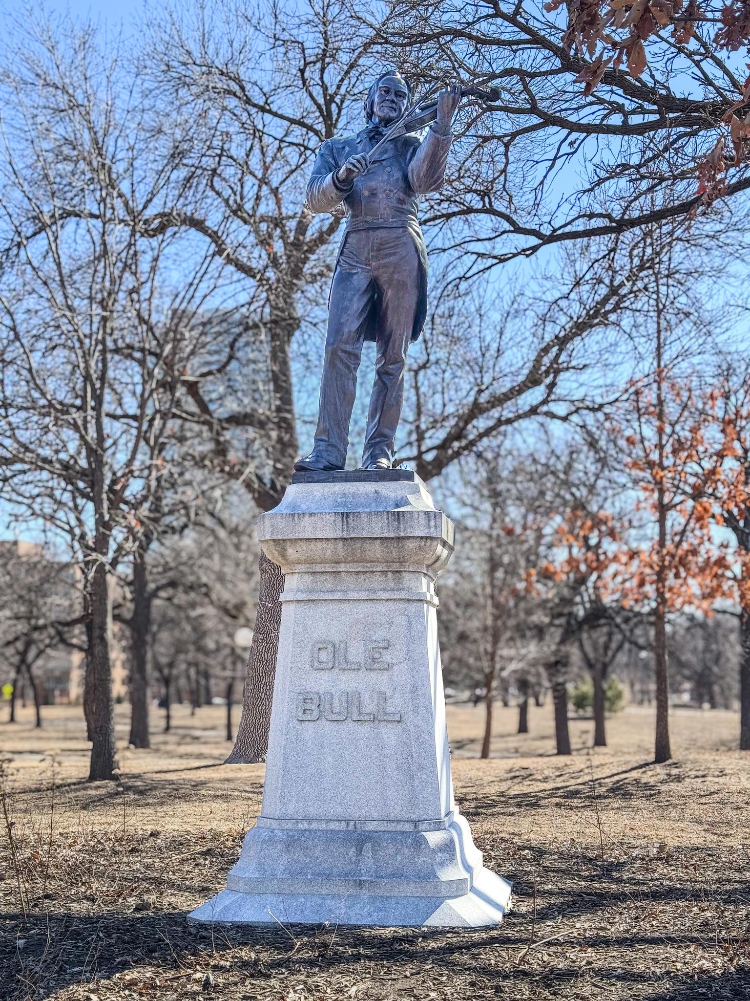Bronze statue of a man on a stone pedestal in a park with bare trees.