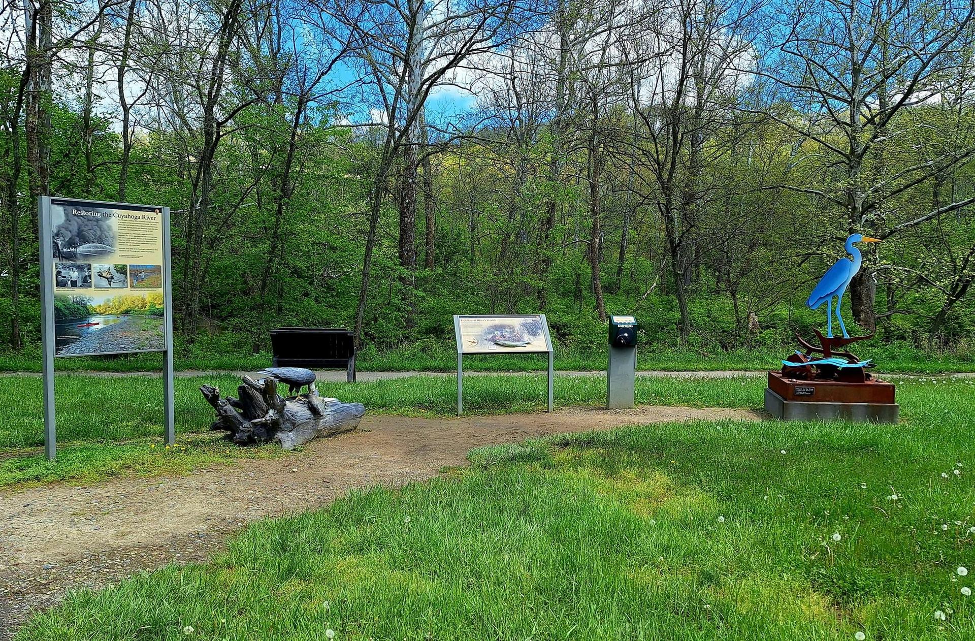 A dirt path leads past information signs and a blue bird sculpture in bright daylight.
