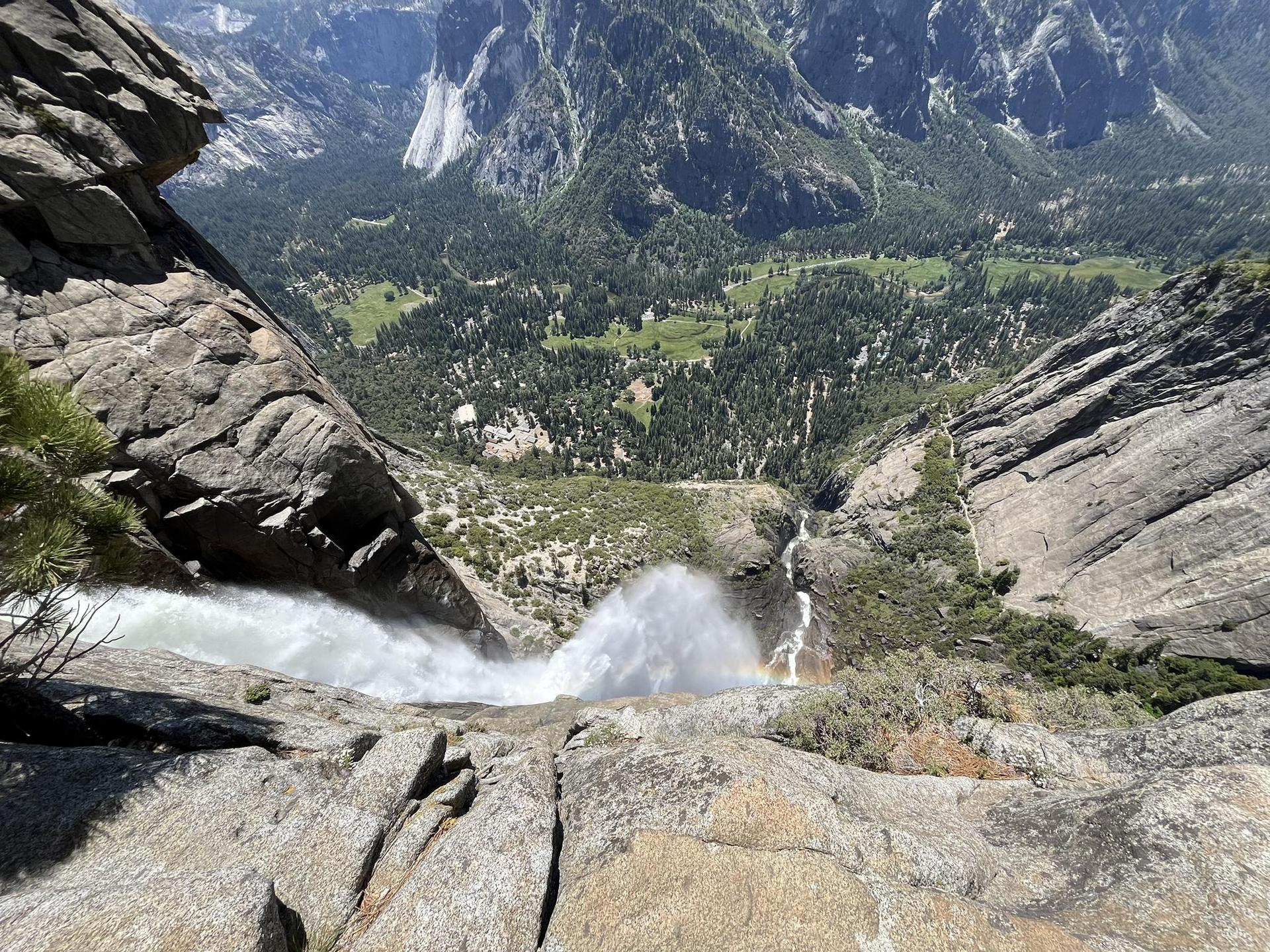 Waterfall plunging into a deep, forested valley from rocky cliffs on a sunny day.