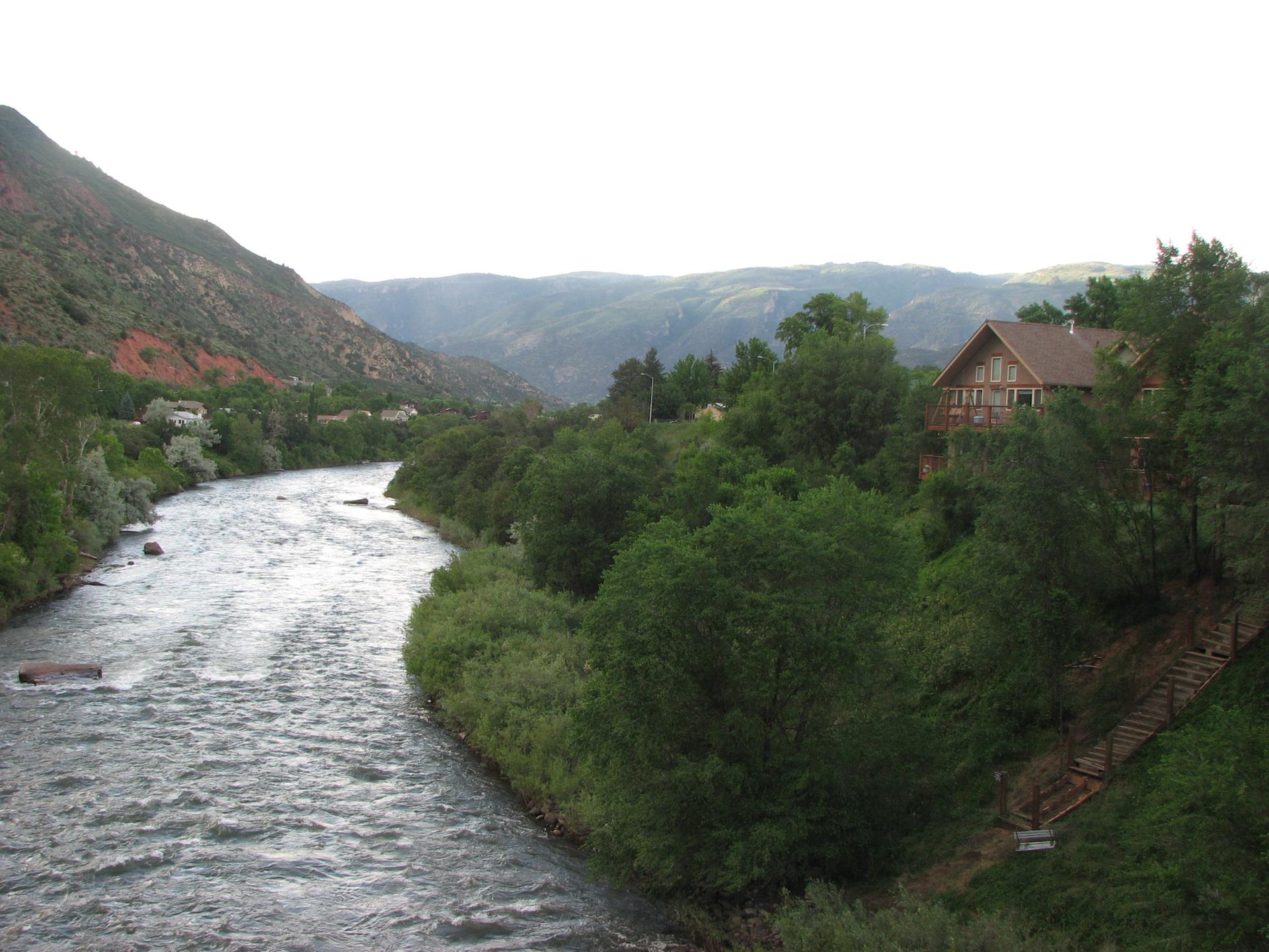 A river flows past a large house on a wooded hillside with distant mountains.