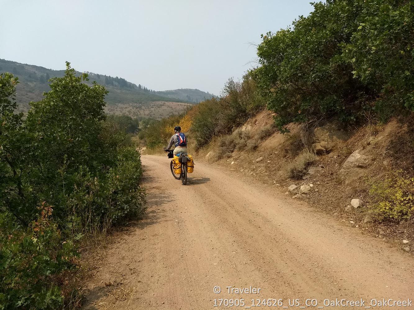 A person cycles away on a dirt road through green, hilly terrain.