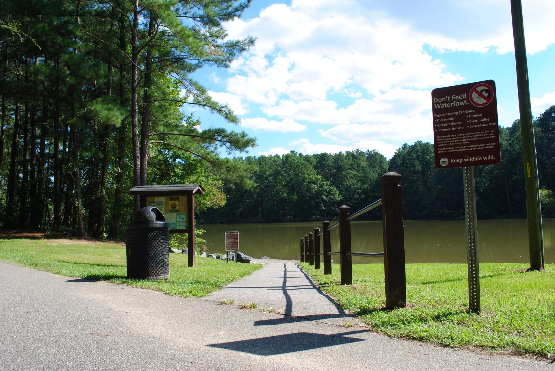A paved path leads to a lake, with signs and a trash can.