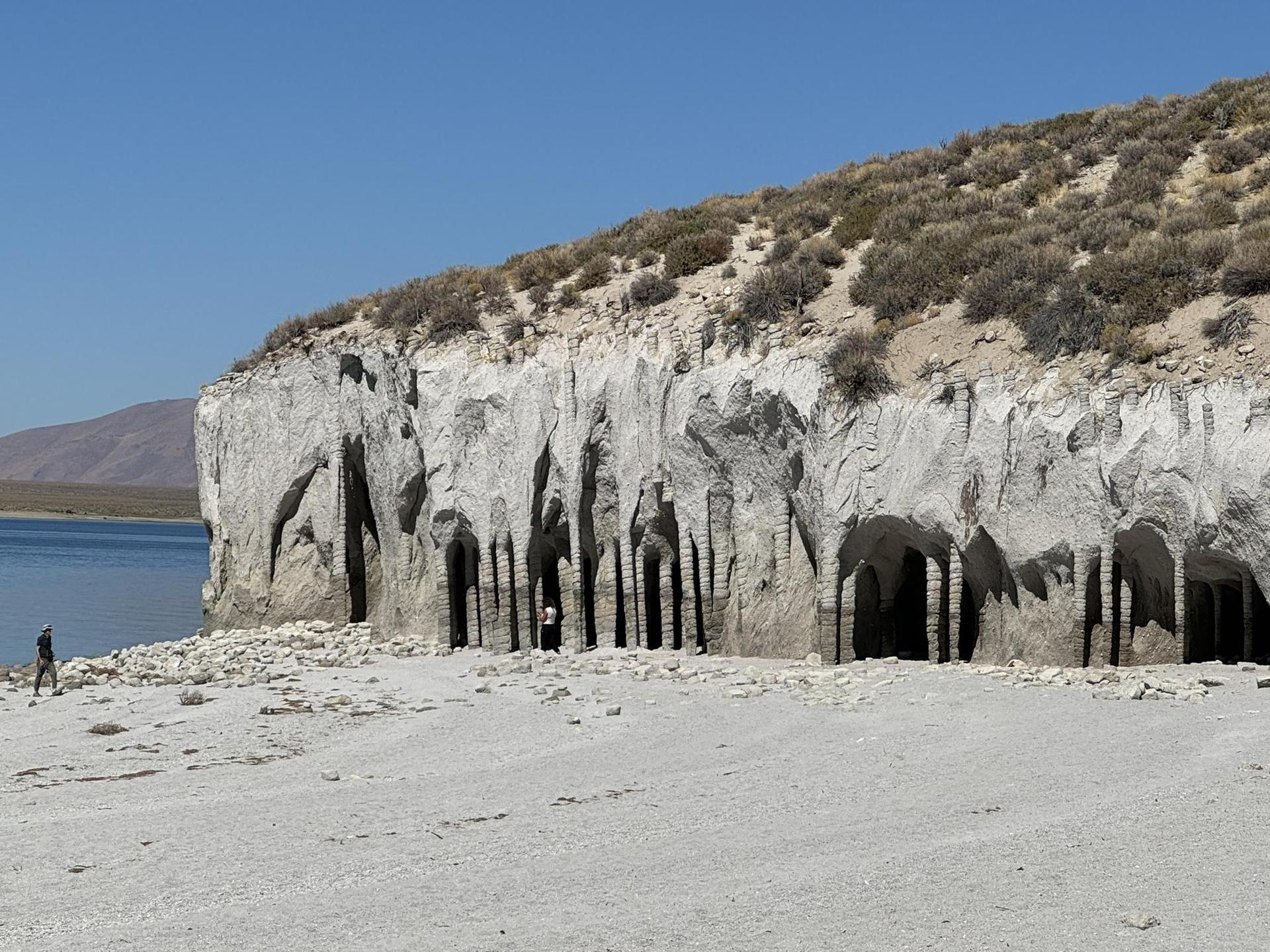 Person walks on a bright shore next to a lake and a cliff with archways.