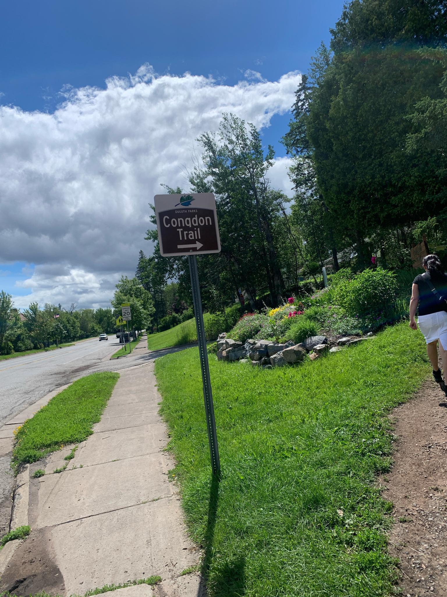 A trail sign next to a sidewalk, green grass, and a person walking.
