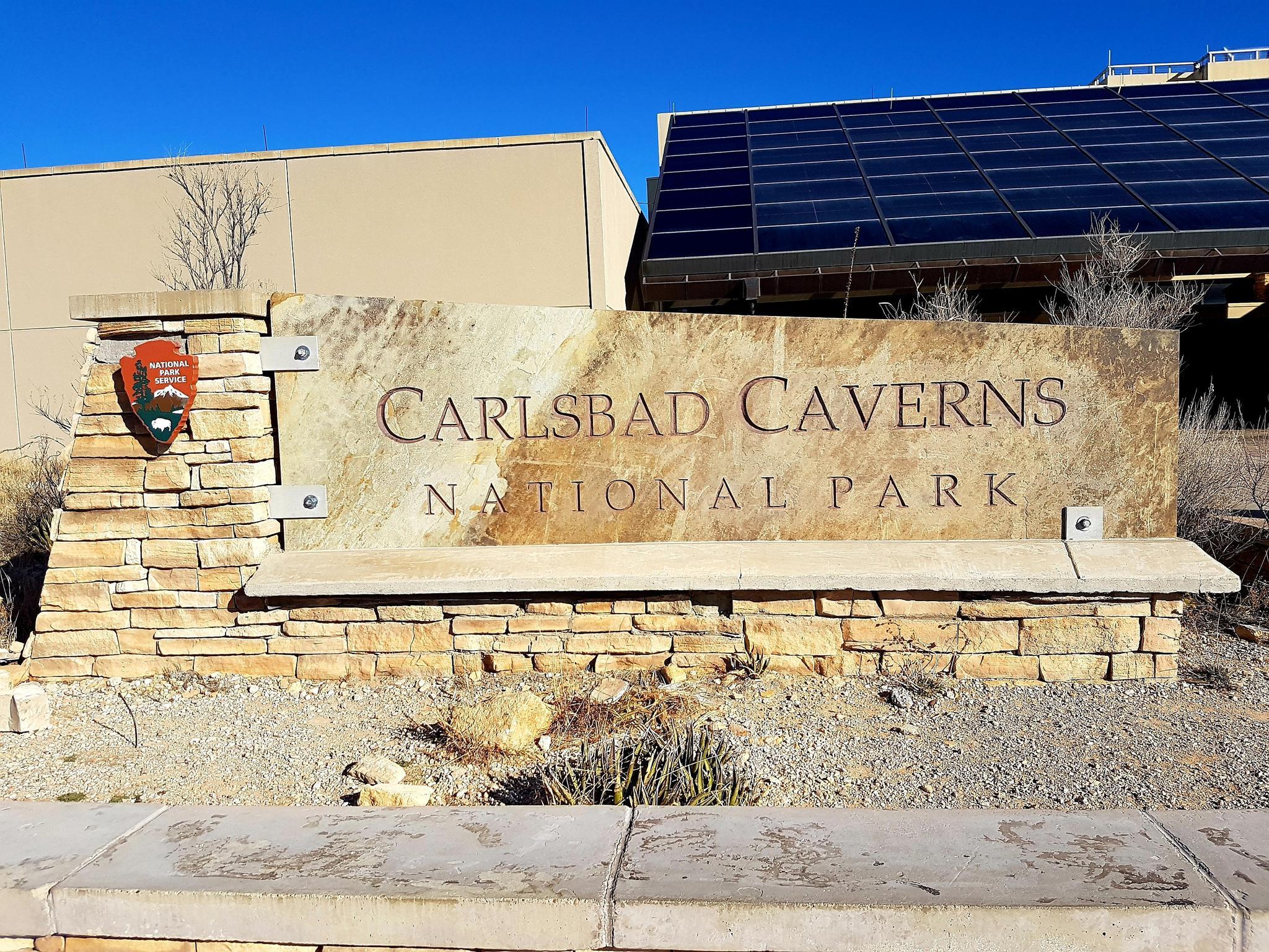 Large stone sign with text, building, and solar panels under bright blue sky.