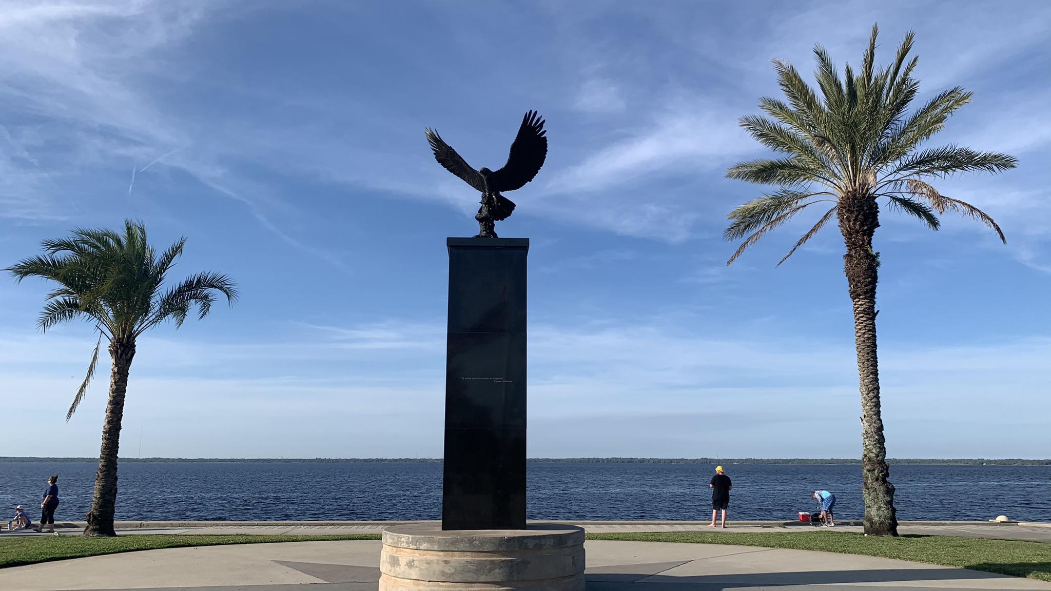 Eagle statue between palm trees overlooking calm water with people.