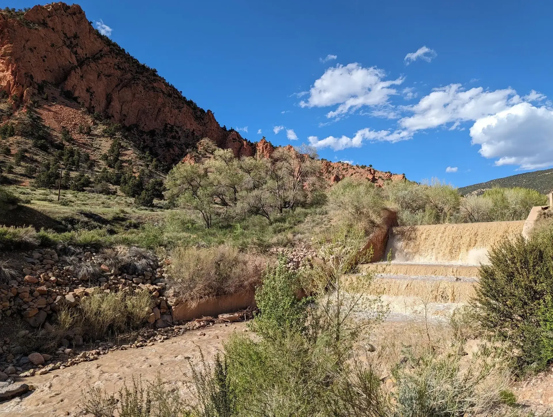 Wide view of a muddy river cascading over a dam in a sunny, rocky canyon.