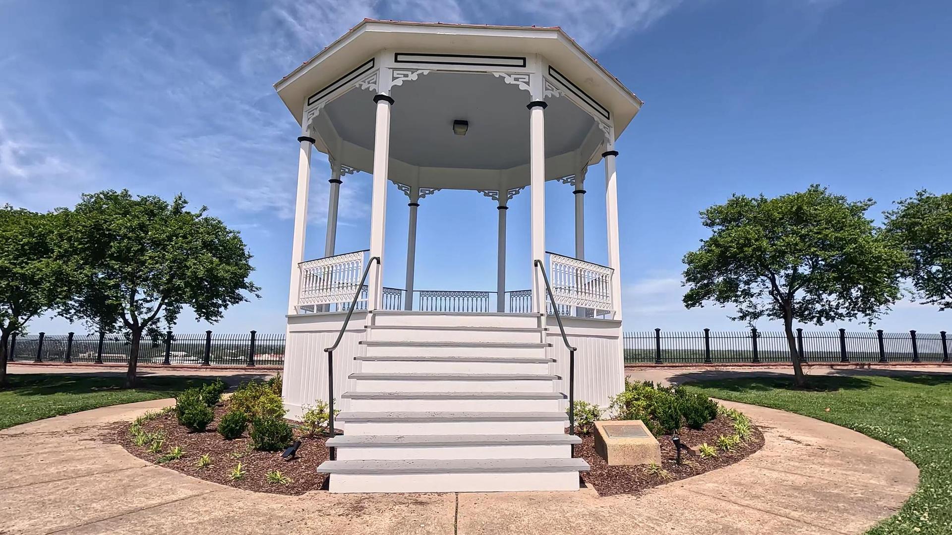 White gazebo with stairs on a sunny day, surrounded by green trees.