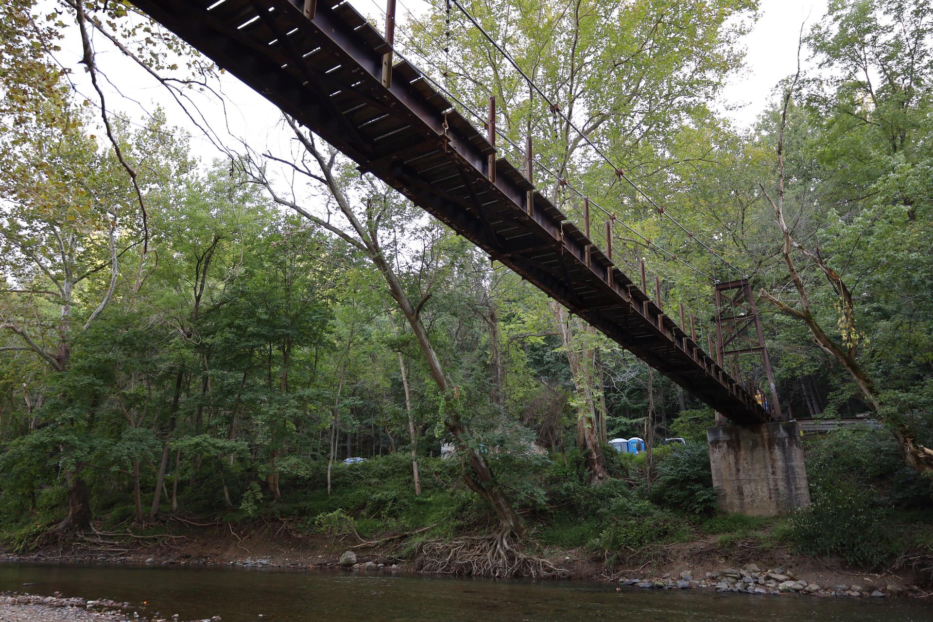 Long wooden suspension bridge spans a river surrounded by green trees.