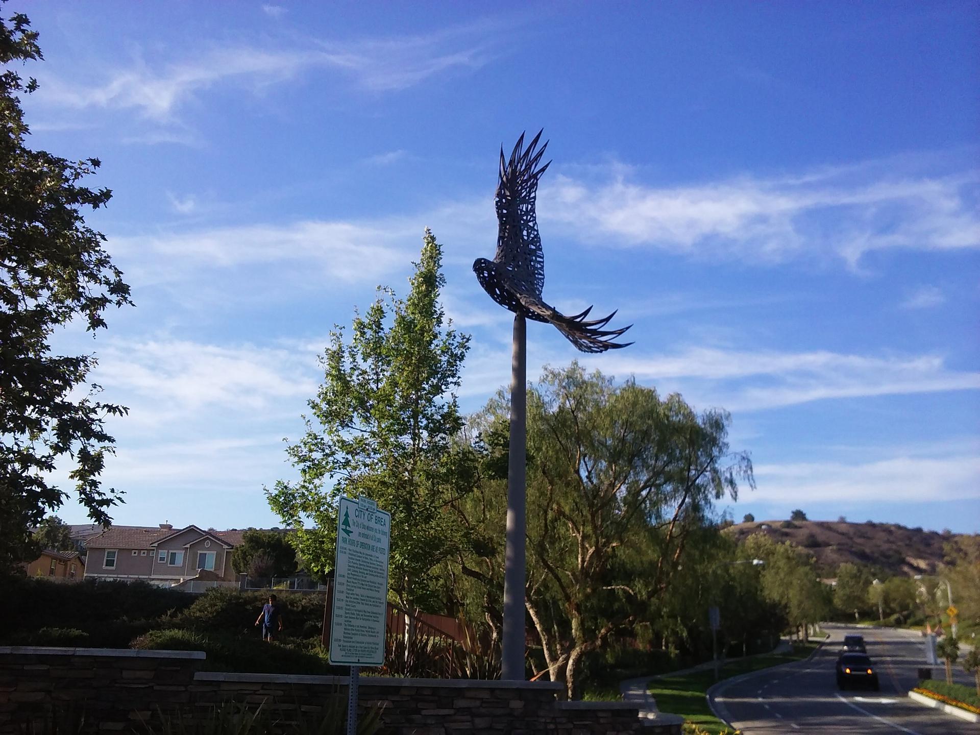A metal bird sculpture on a pole against a bright blue sky with trees.