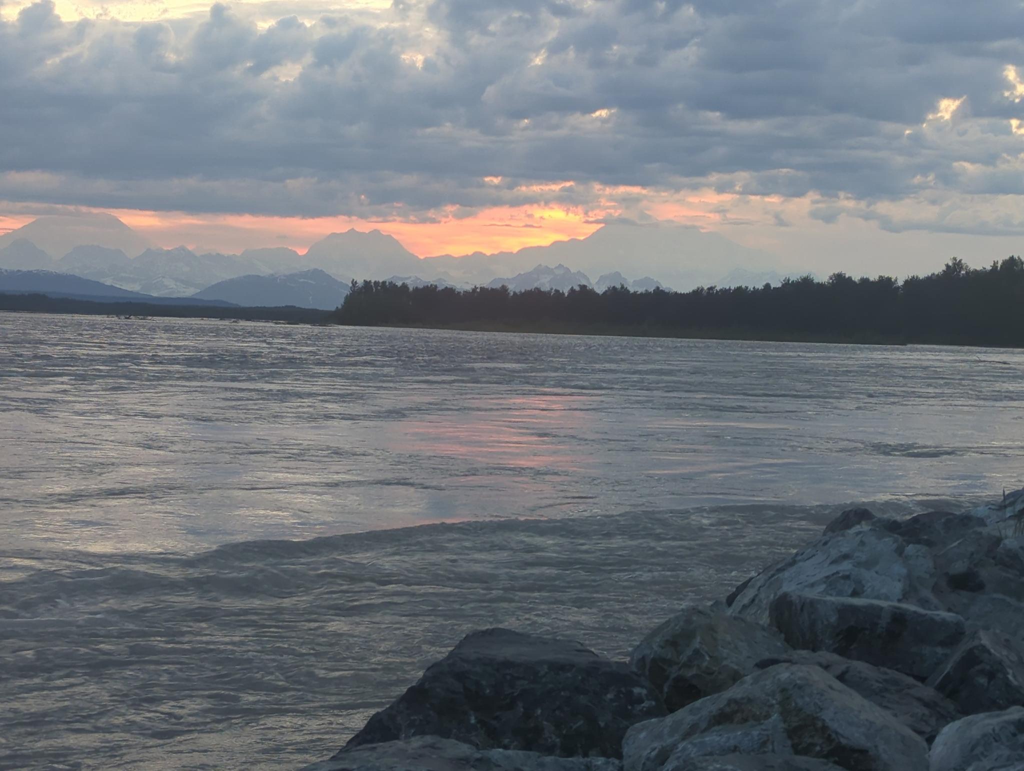 Sunset over a wide body of water with rocky foreground and distant mountains.