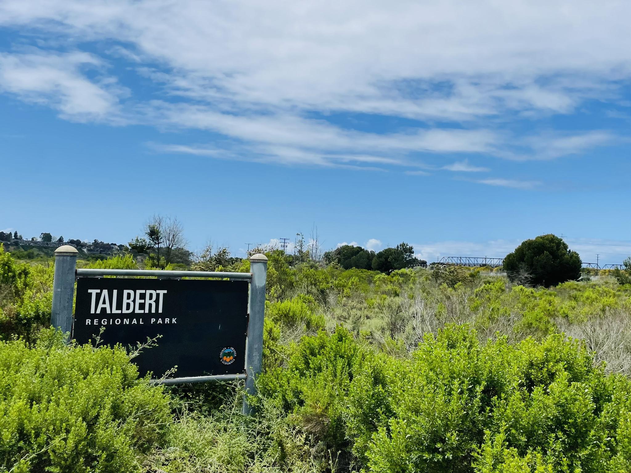 A black sign stands amidst green bushes under a bright, partly cloudy sky.