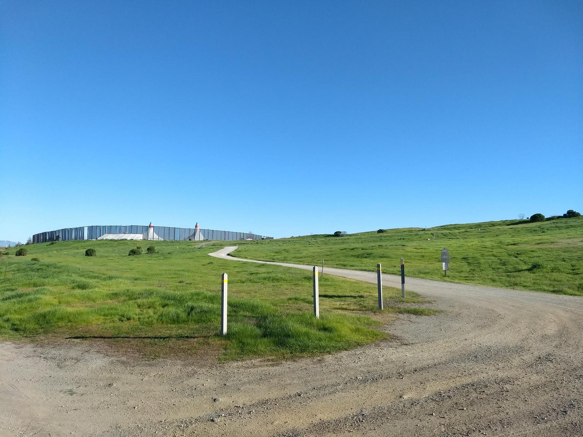 Dirt path curving through green hills towards a distant structure under blue sky.