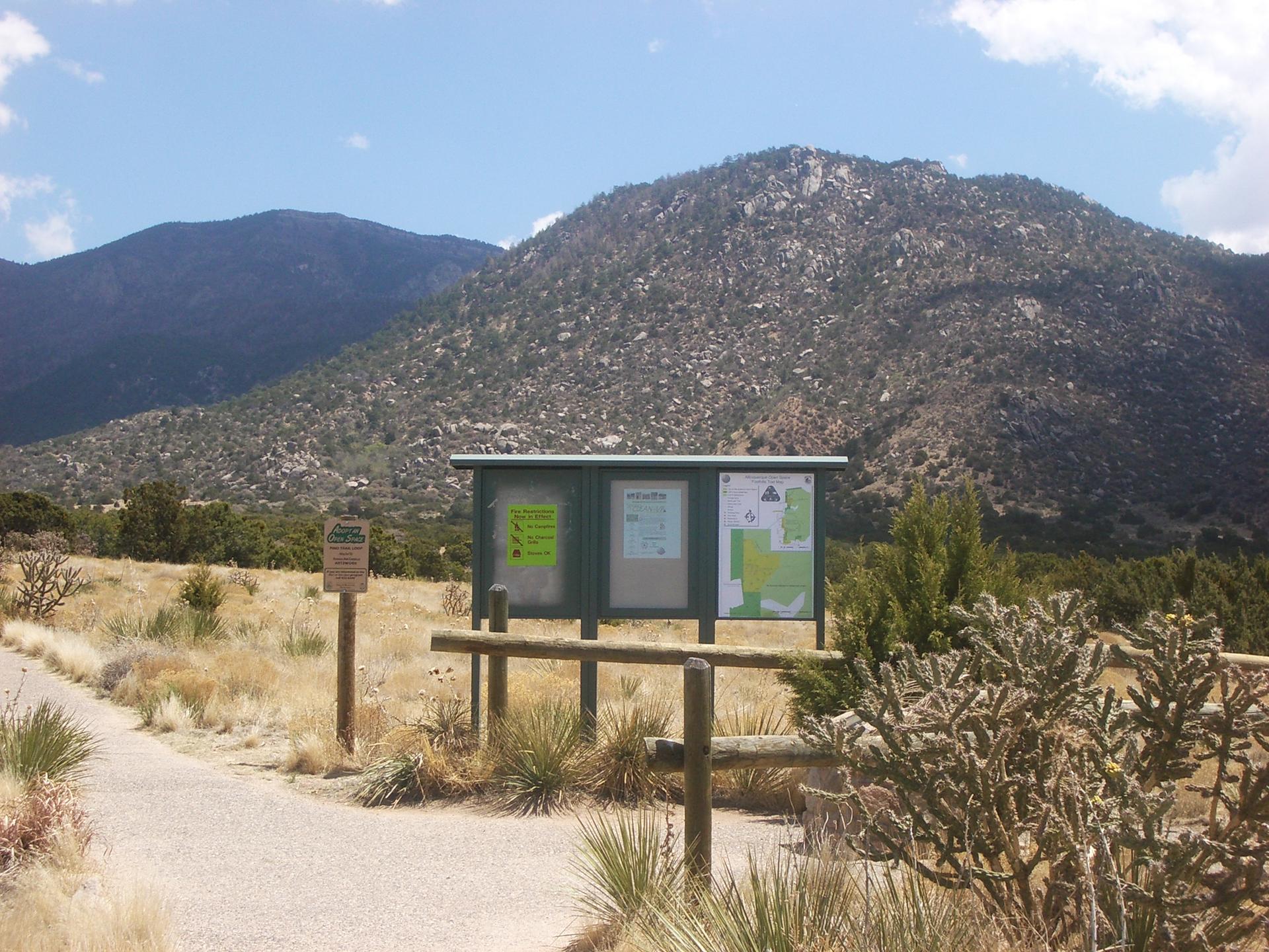 Information board in a grassy field with large, rocky mountains under a bright sky.
