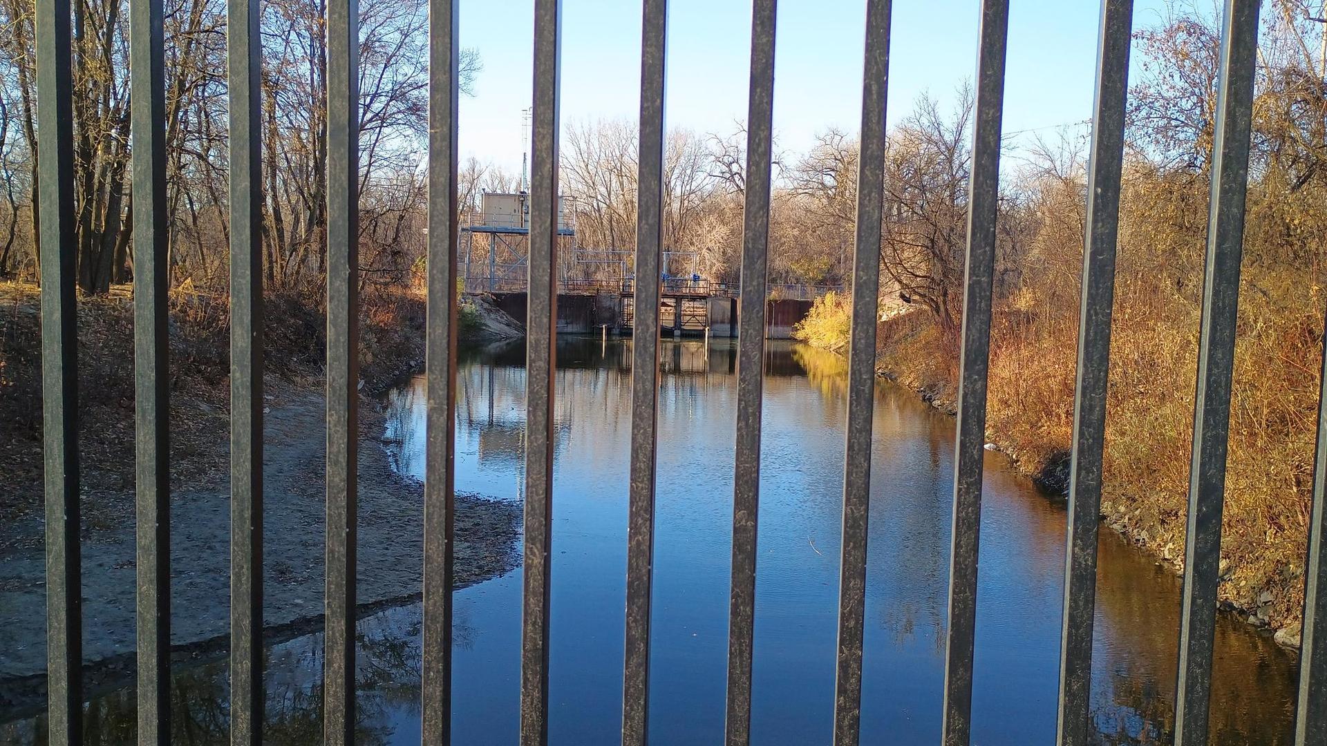 View through a metal fence of a calm river and distant trees.