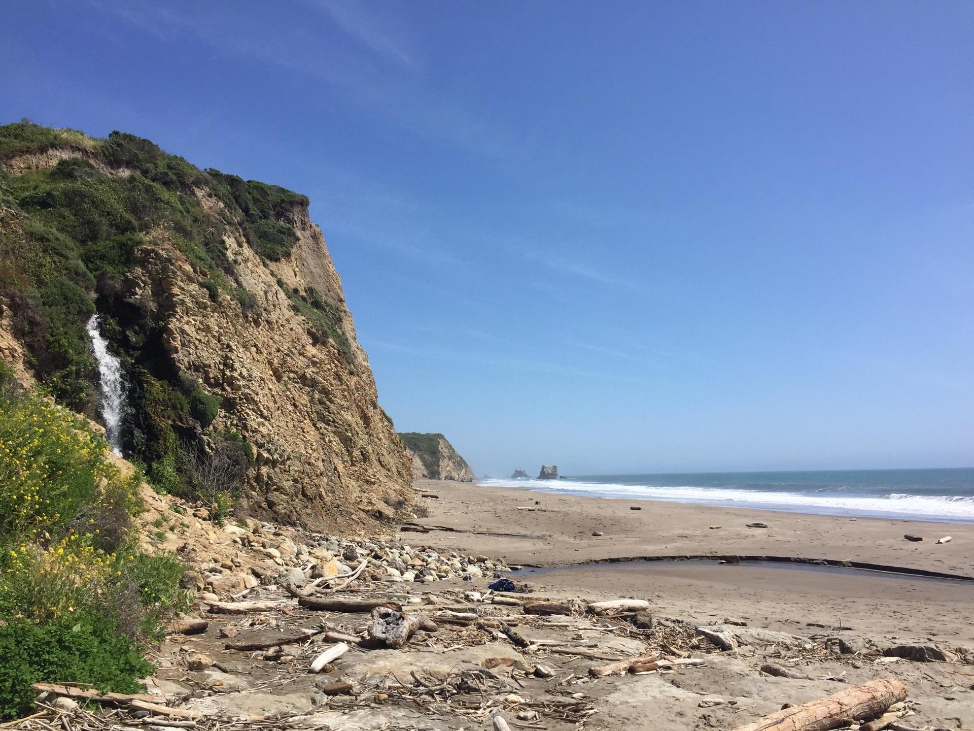 A small waterfall cascades down a tall cliff onto a wide sandy beach.