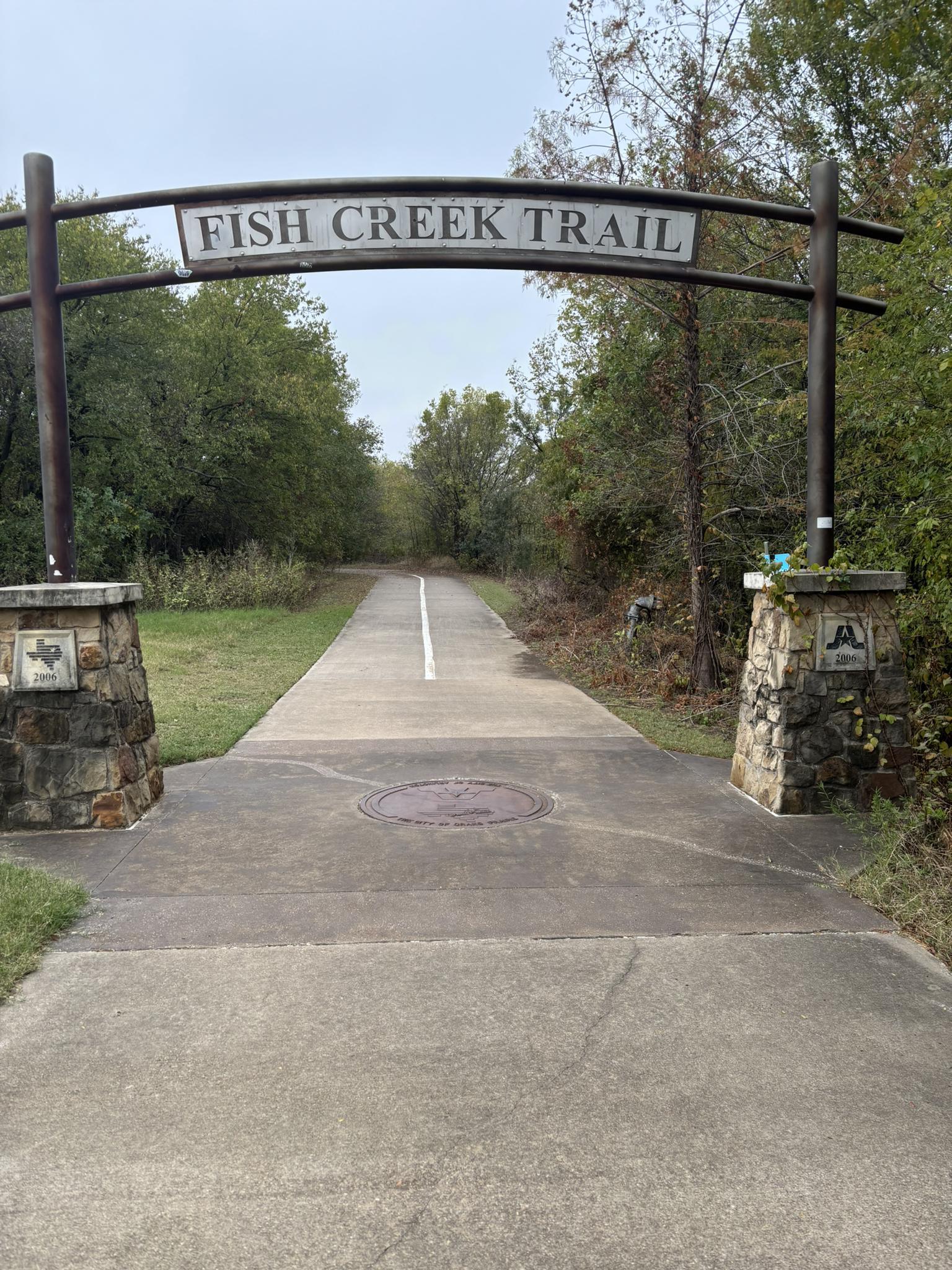 Paved trail entrance with a decorative archway, leading into a wooded path.