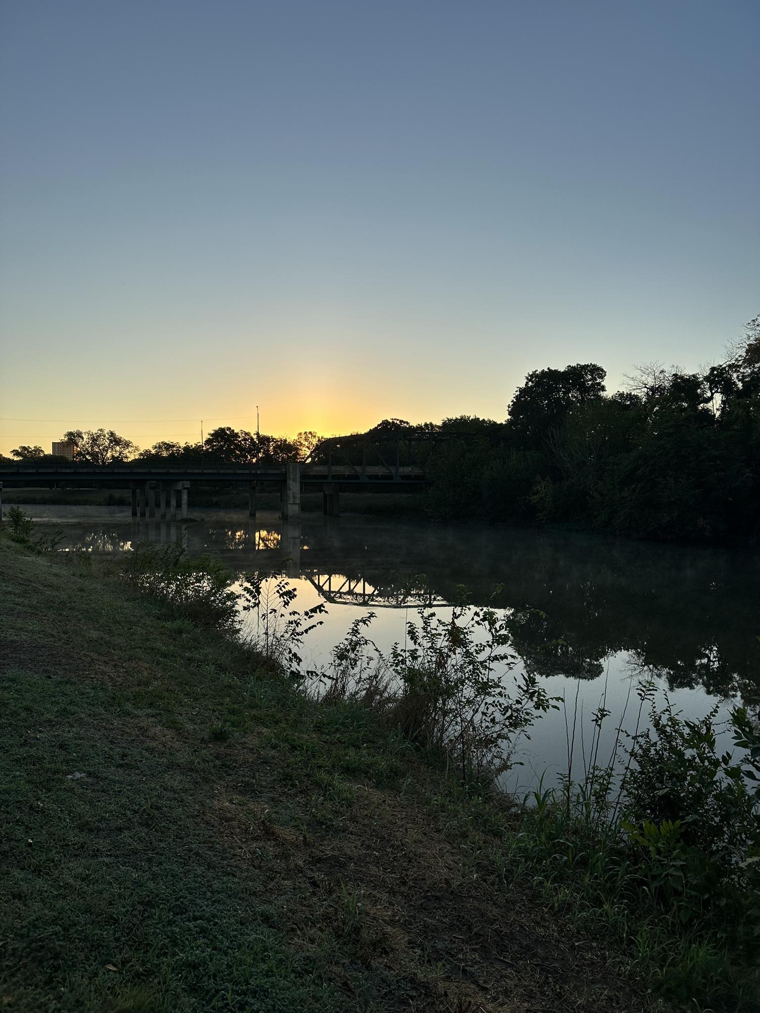 Sunset over a river with a bridge and trees silhouetted against the sky.