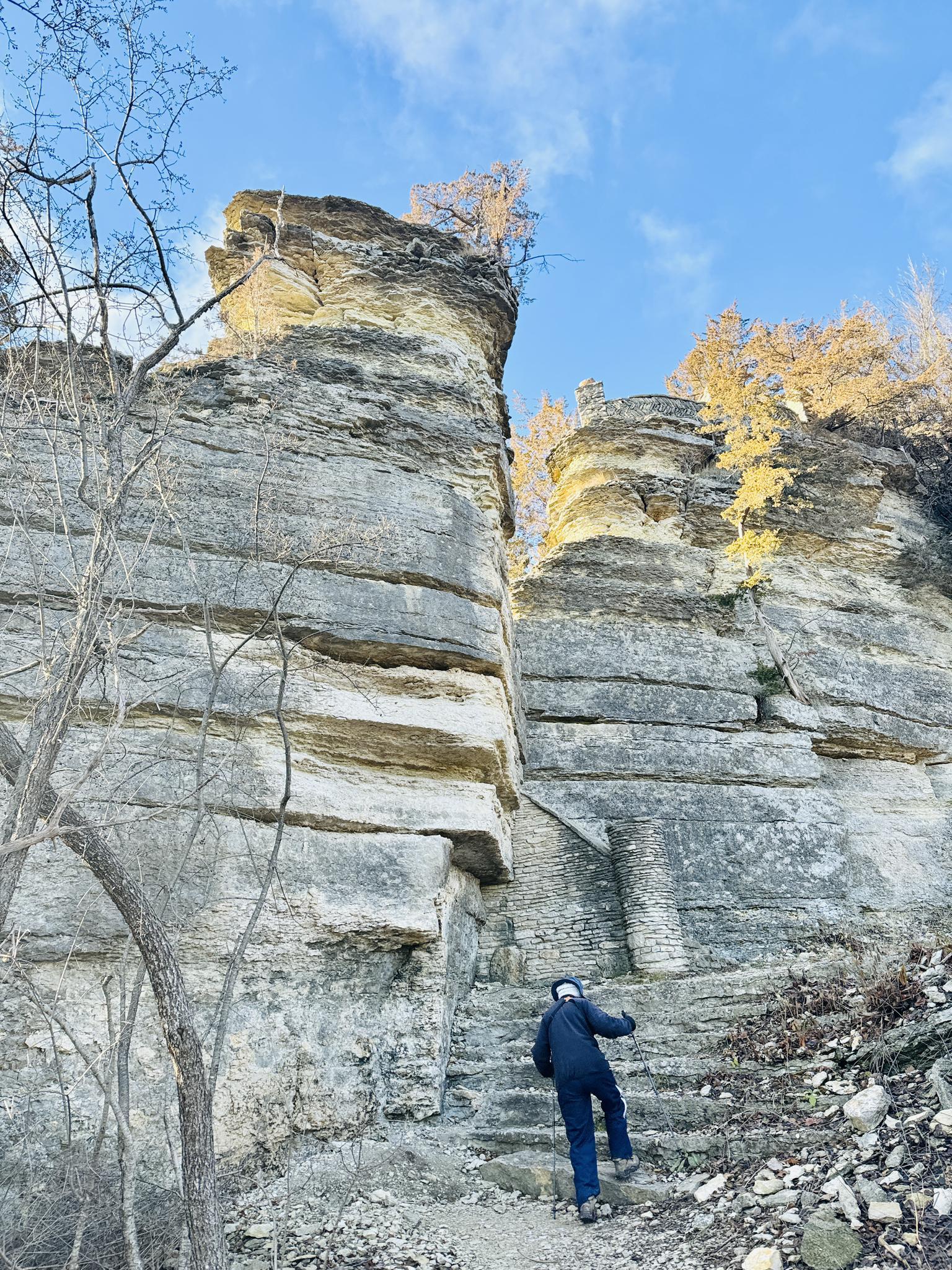 Person climbs stone steps beside tall, layered rock formations under a bright blue sky.
