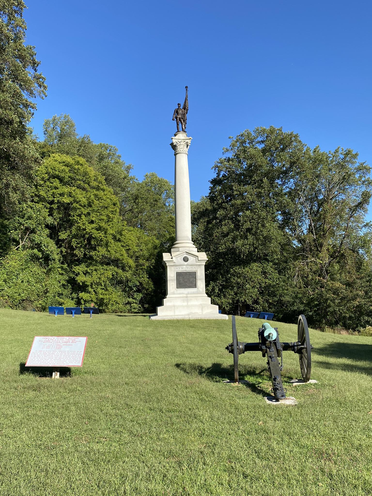 Tall monument with a statue, cannon, and sign in a grassy field.