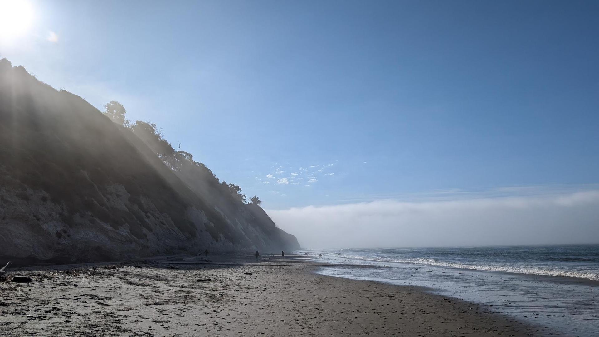 Sunlit misty beach with a steep cliff and distant figures walking.