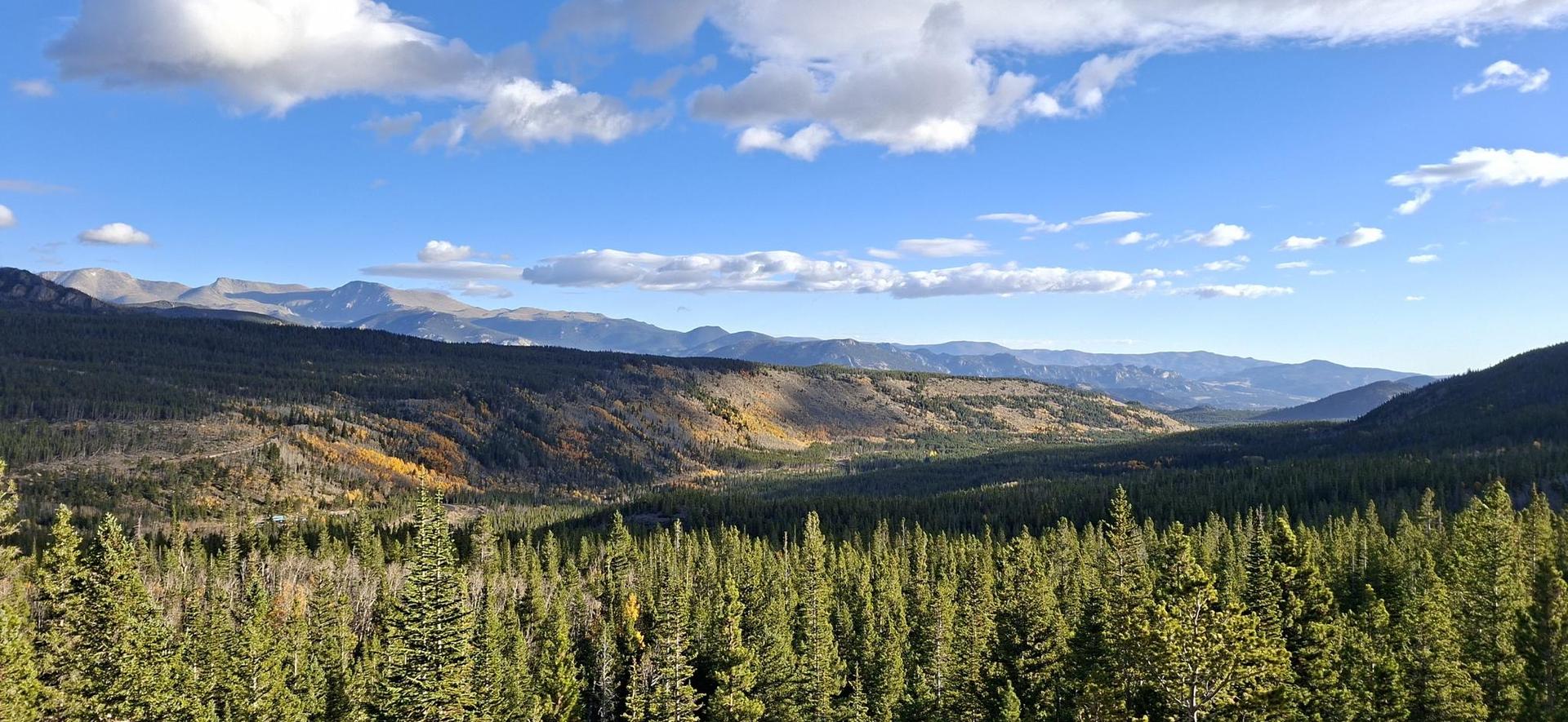Expansive mountain landscape with dense forests and distant peaks under blue sky.