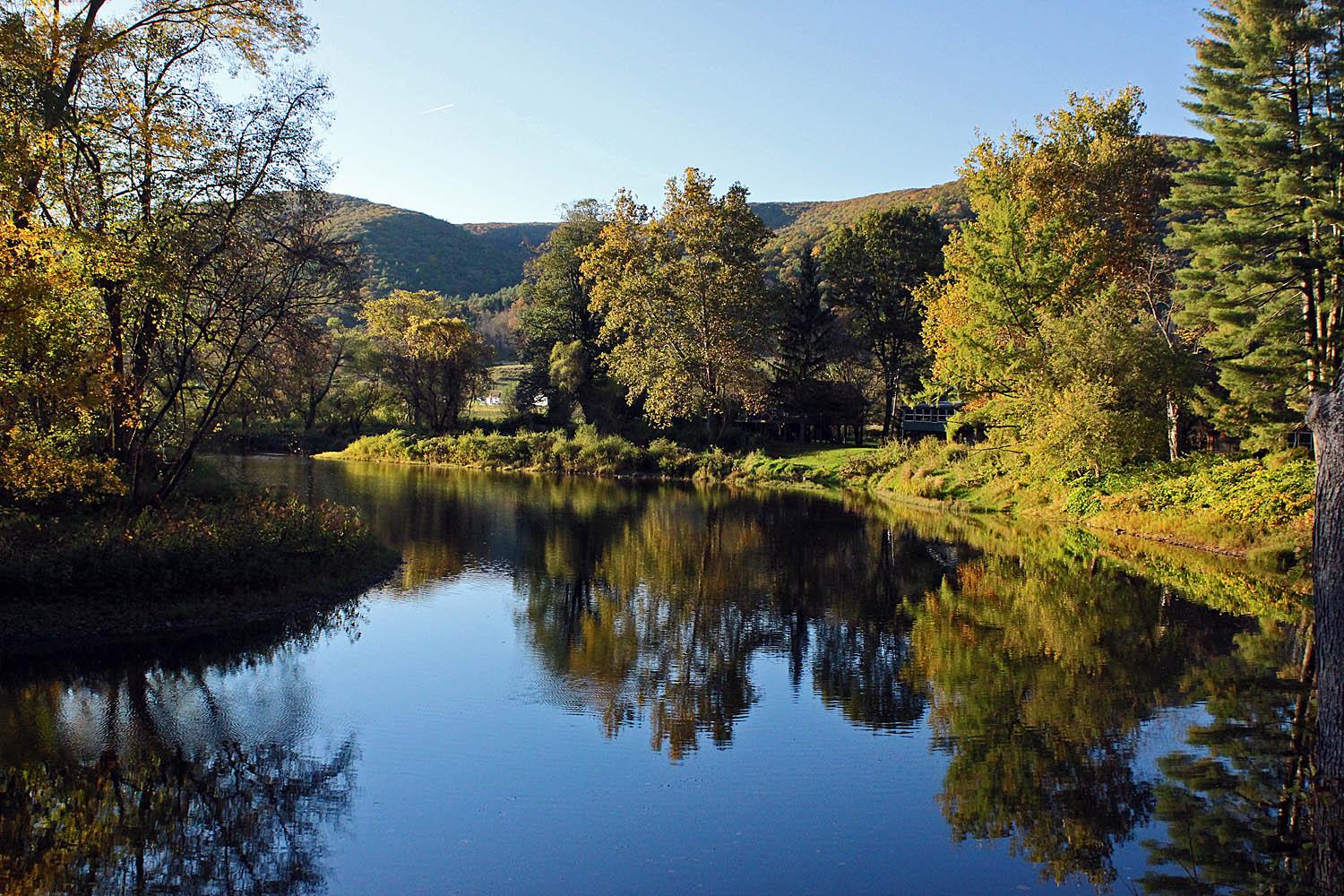 Calm river reflecting autumn trees and distant hills on a clear day.