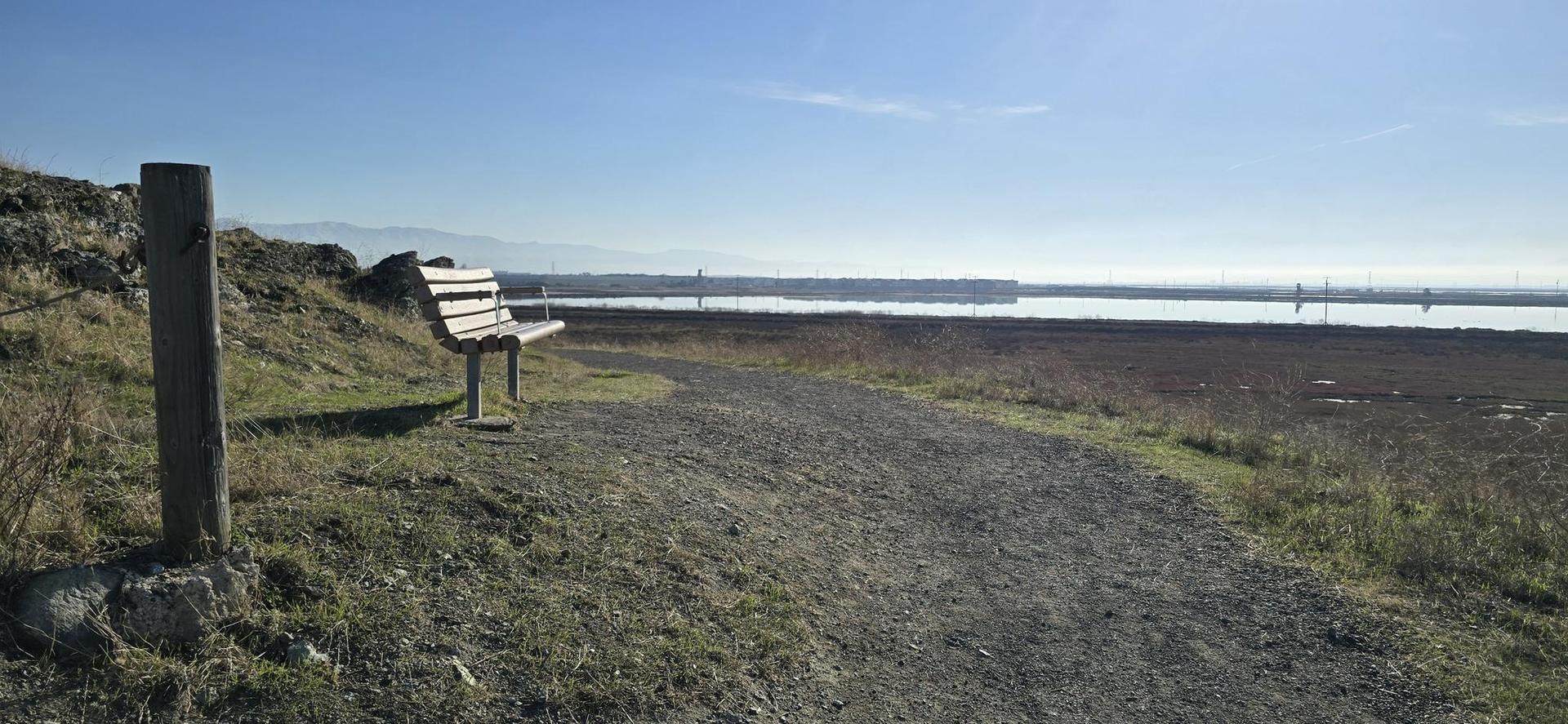 An empty wooden bench sits beside a gravel path overlooking water.