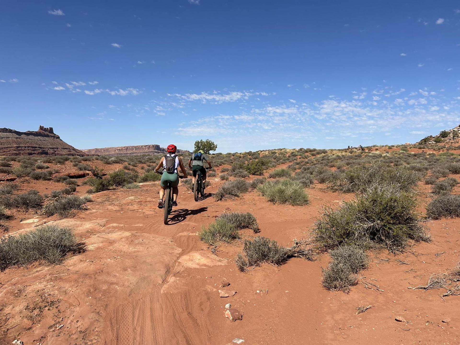 Two people mountain biking on a red dirt trail under a clear blue sky.