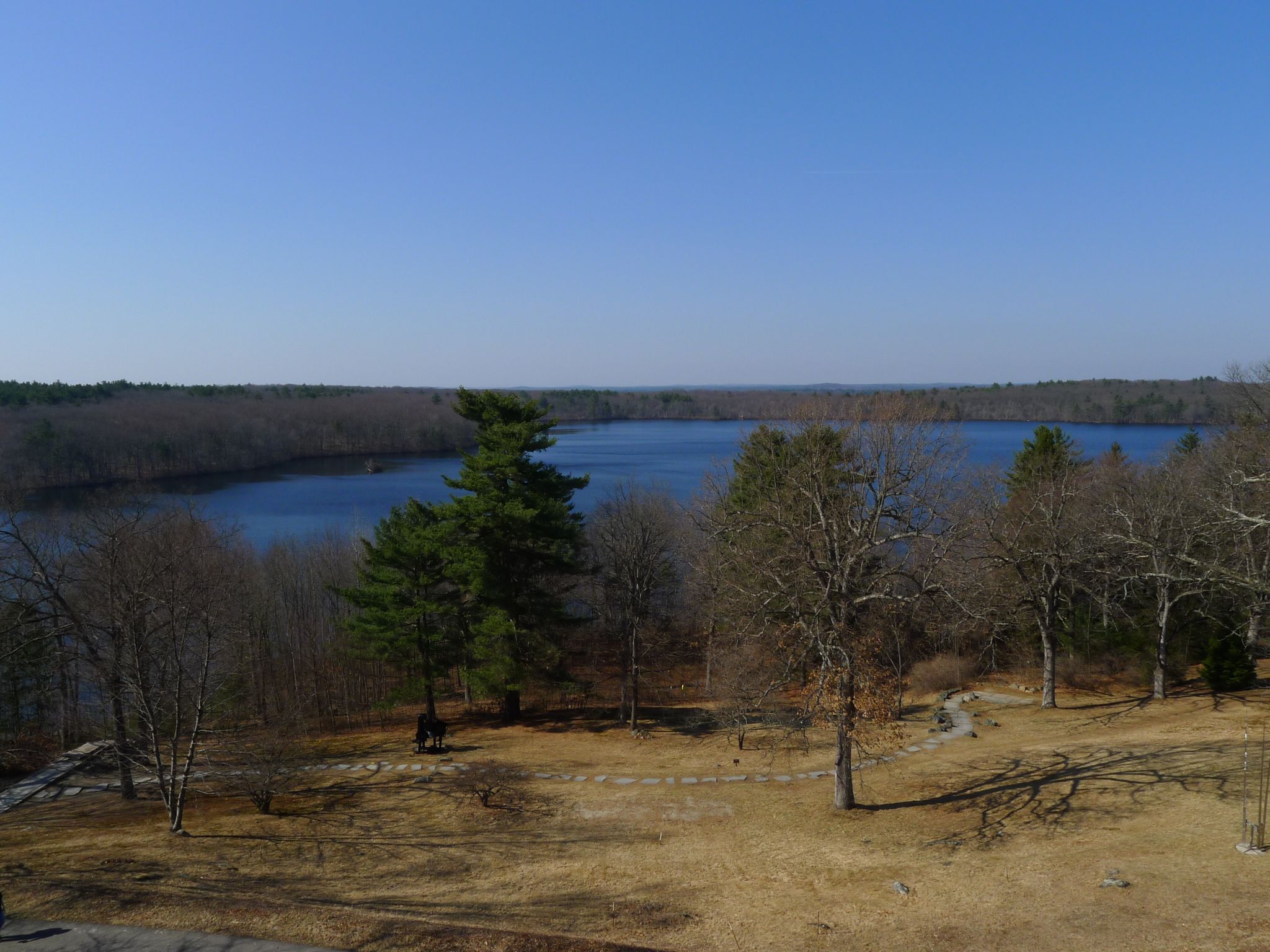 Wide view of a lake surrounded by trees under a clear blue sky. - Flints Pond Trail