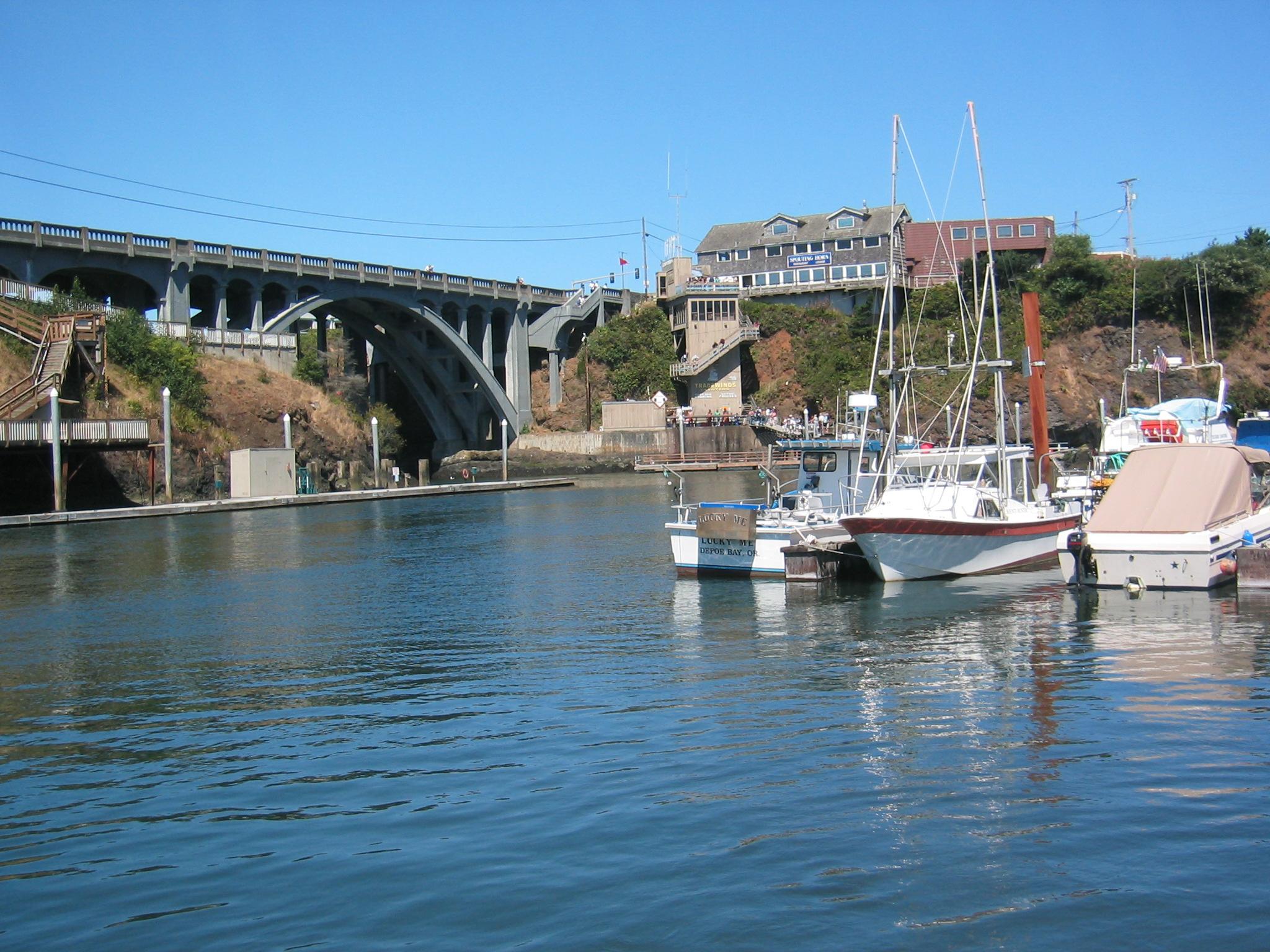 Sunny view of boats docked in a harbor under a large arched bridge.