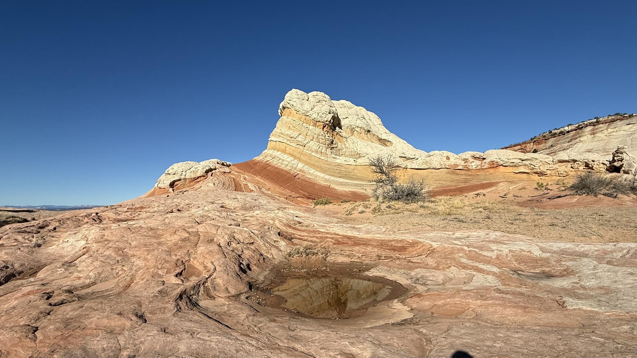 Layered white and red rock formation with a small water pool under a clear blue sky.