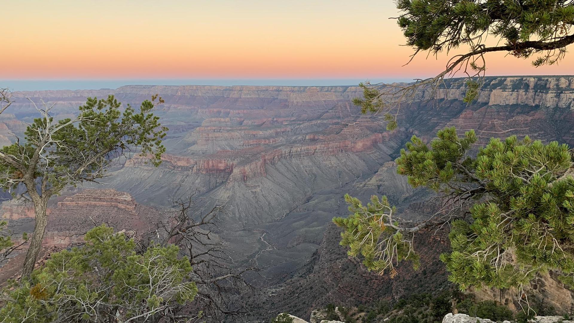 Canyon landscape at sunset with colorful sky, framed by green trees.