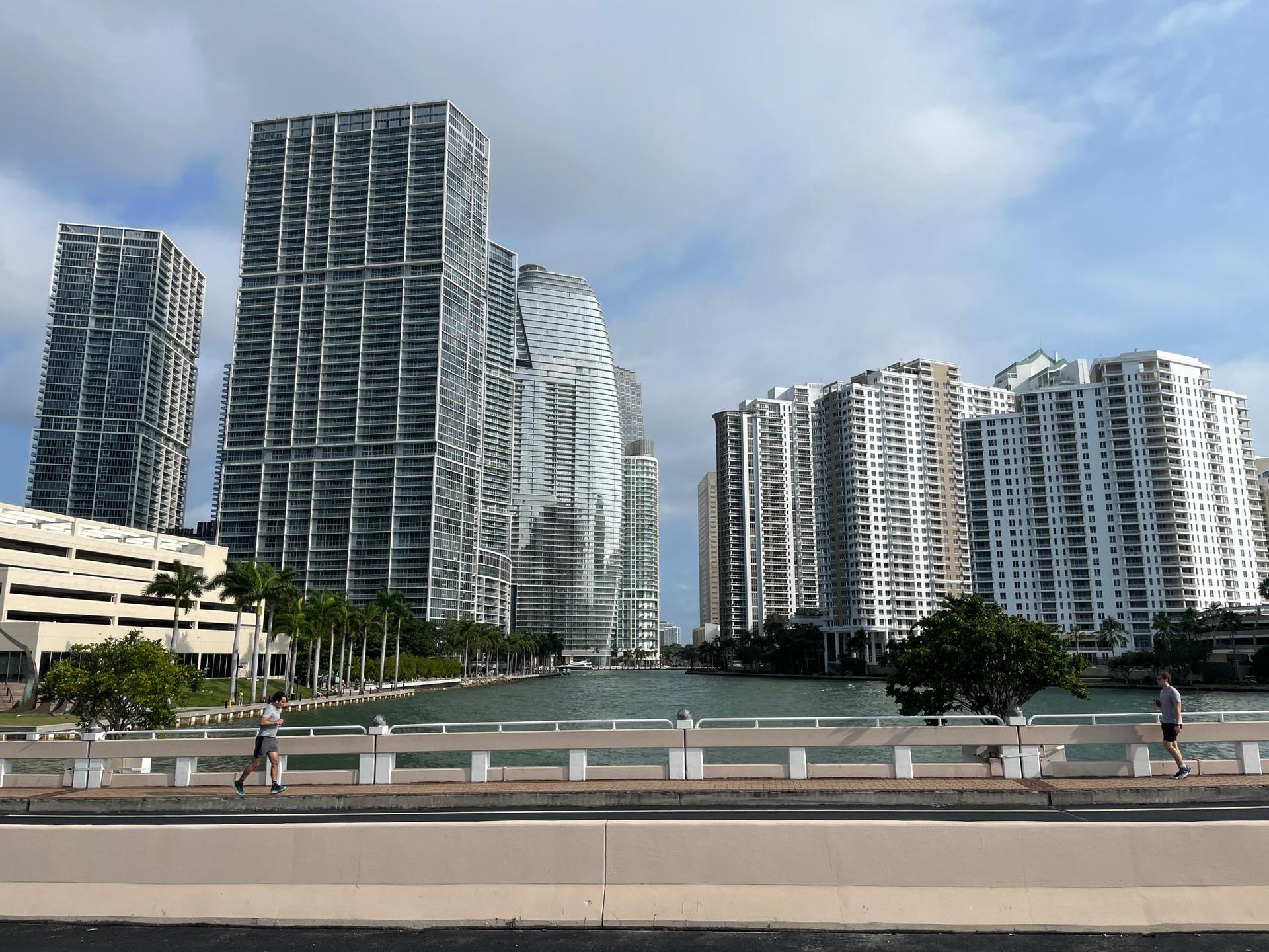 Two people walk on a bridge overlooking a city skyline and water.
